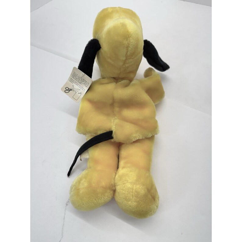 Puede incluir: Un juguete de peluche con forma de perro amarillo con orejas y cola negras. El juguete tiene una etiqueta adjunta. El perro est&aacute; en posici&oacute;n sentada, con las patas extendidas. El juguete es de material suave.