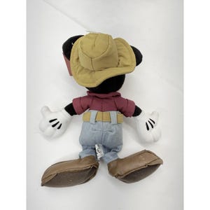 K&ouml;nnte beinhalten: Ein Pl&uuml;schtier von Mickey Mouse, von hinten gesehen, mit einem beigefarbenen Cowboyhut, einem weinroten Hemd, einer blauen Jeans und braunen Stiefeln. Das Spielzeug hat wei&szlig;e Handschuhe mit schwarzen Linien und schwarze Ohren. Das Spielzeug befindet sich auf wei&szlig;em Hintergrund.