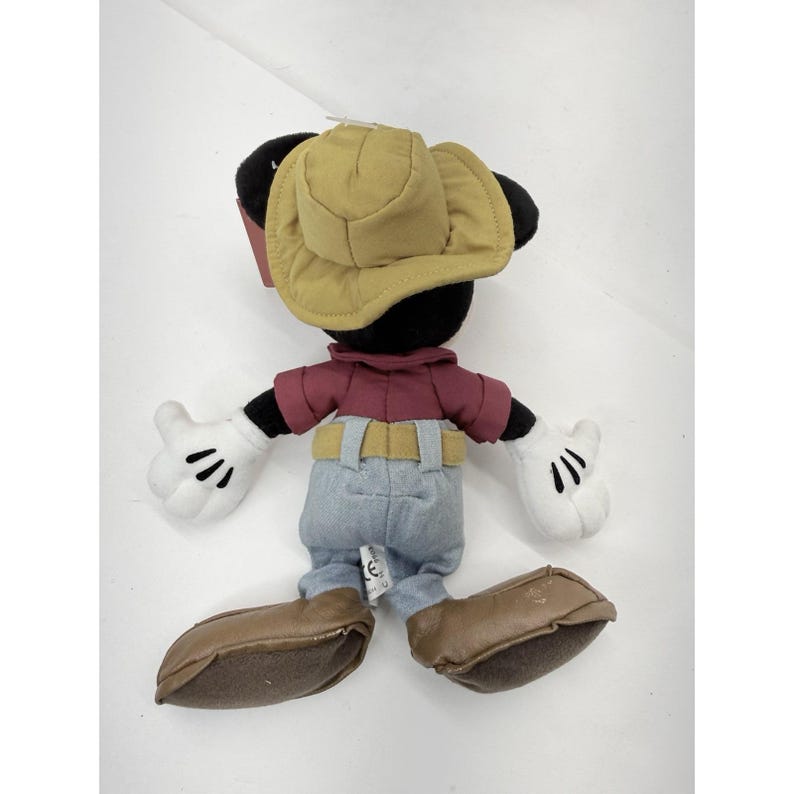 K&ouml;nnte beinhalten: Ein Pl&uuml;schtier von Mickey Mouse, von hinten gesehen, mit einem beigefarbenen Cowboyhut, einem weinroten Hemd, einer blauen Jeans und braunen Stiefeln. Das Spielzeug hat wei&szlig;e Handschuhe mit schwarzen Linien und schwarze Ohren. Das Spielzeug befindet sich auf wei&szlig;em Hintergrund.