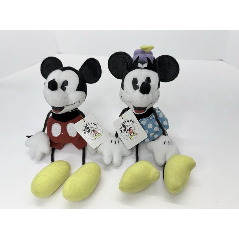 K&ouml;nnte beinhalten: Zwei Mickey Mouse Pl&uuml;schtiere mit schwarzen Ohren, wei&szlig;en Handschuhen und gelben F&uuml;&szlig;en. Einer tr&auml;gt ein rotes Outfit, der andere ein blau gepunktetes Kleid. Jeder hat ein "Mickey & Co." Etikett.