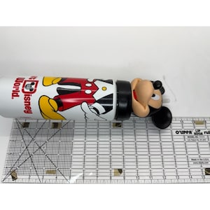 May include: A white Disney World water bottle featuring Mickey Mouse in his classic outfit. The bottle has a black cap and a Mickey Mouse head on the end. The bottle is approximately 10 inches long.