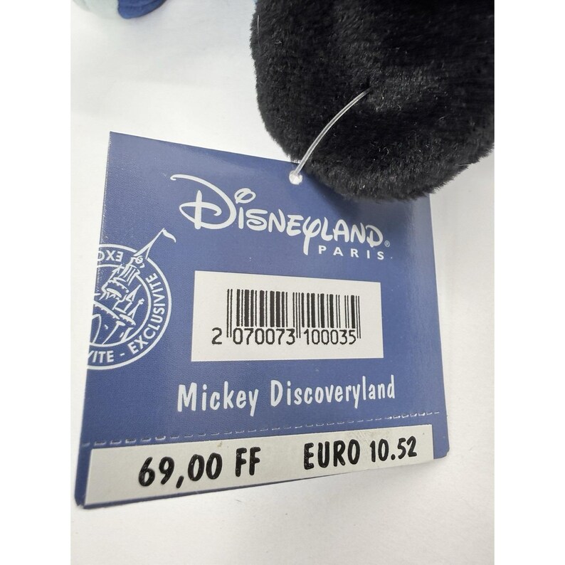 May include: A black plush toy with a blue and white accent. A blue tag is attached, featuring the Disneyland Paris logo and the text "Mickey Discoveryland". The tag also includes a barcode.
