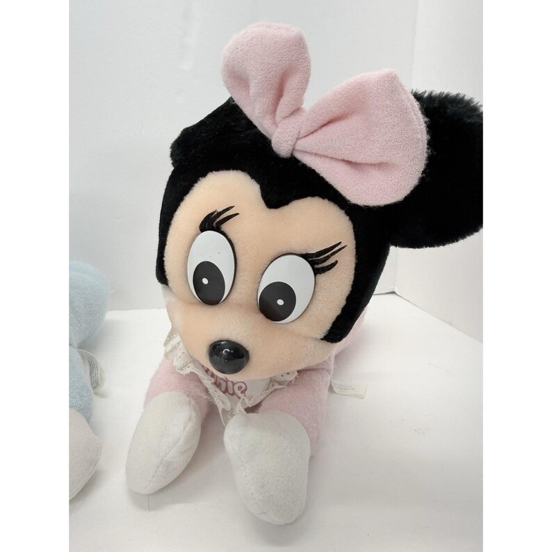 Puede incluir: Mu&ntilde;eca de peluche de Minnie Mouse con un lazo rosa, orejas negras y un atuendo rosa y blanco. La mu&ntilde;eca tiene ojos grandes y expresivos y un cuello de encaje. La mu&ntilde;eca est&aacute; acostada.