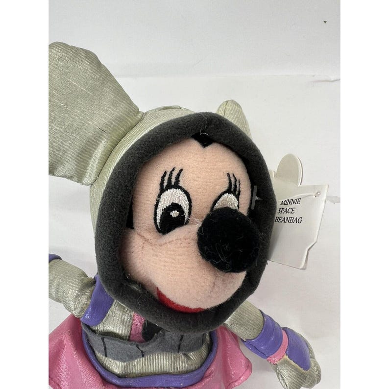 May include: A plush Minnie Mouse beanbag toy wearing a silver space suit with a pink and purple cape. The tag reads "Minnie Space Beanbag".