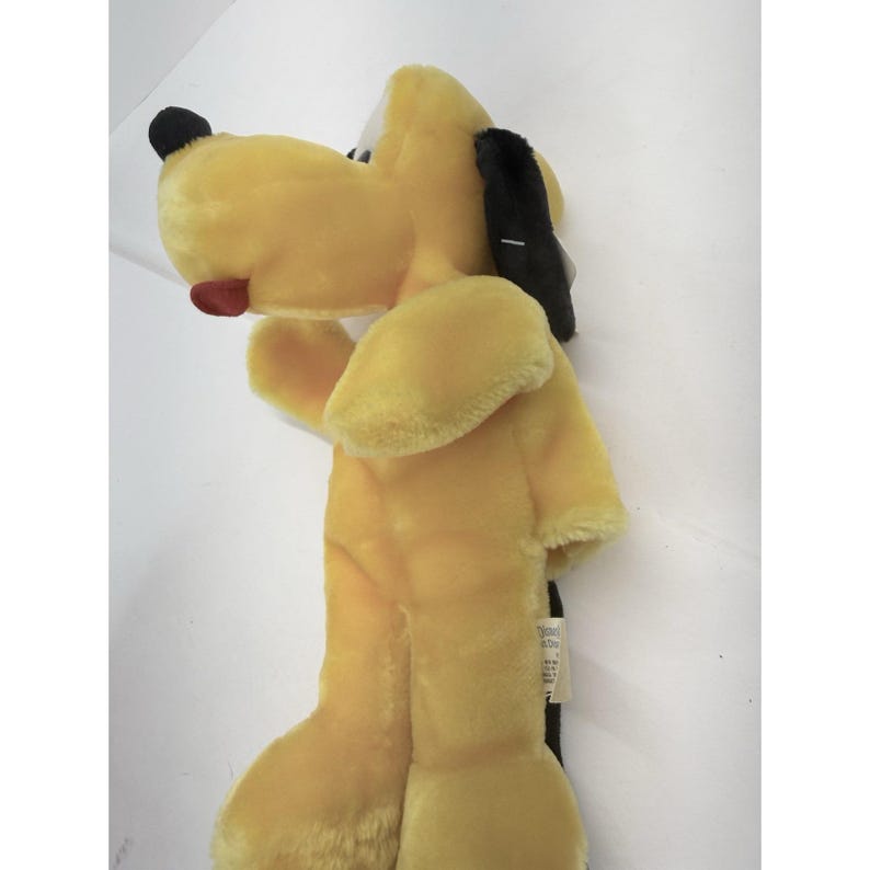 Puede incluir: Un peluche de Pluto, un perro amarillo con orejas y nariz negras. El juguete tiene una lengua roja y una mancha negra en la cabeza. El juguete est&aacute; hecho de material suave y afelpado y est&aacute; de pie.