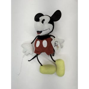K&ouml;nnte beinhalten: Ein Mickey Mouse Pl&uuml;schtier mit schwarzem Kopf und Ohren, wei&szlig;em Gesicht und H&auml;nden, roter Hose mit wei&szlig;en Kn&ouml;pfen und gelben Schuhen. Das Spielzeug hat ein Etikett mit der Aufschrift "Mickey & Co."