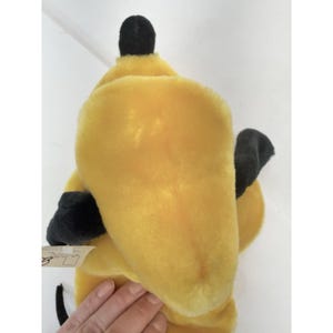 Puede incluir: Un juguete de peluche amarillo con detalles negros, incluyendo una parte superior y orejas negras. El juguete tiene una textura suave y una etiqueta adjunta. Es probable que sea un personaje de un cuento infantil o de dibujos animados.