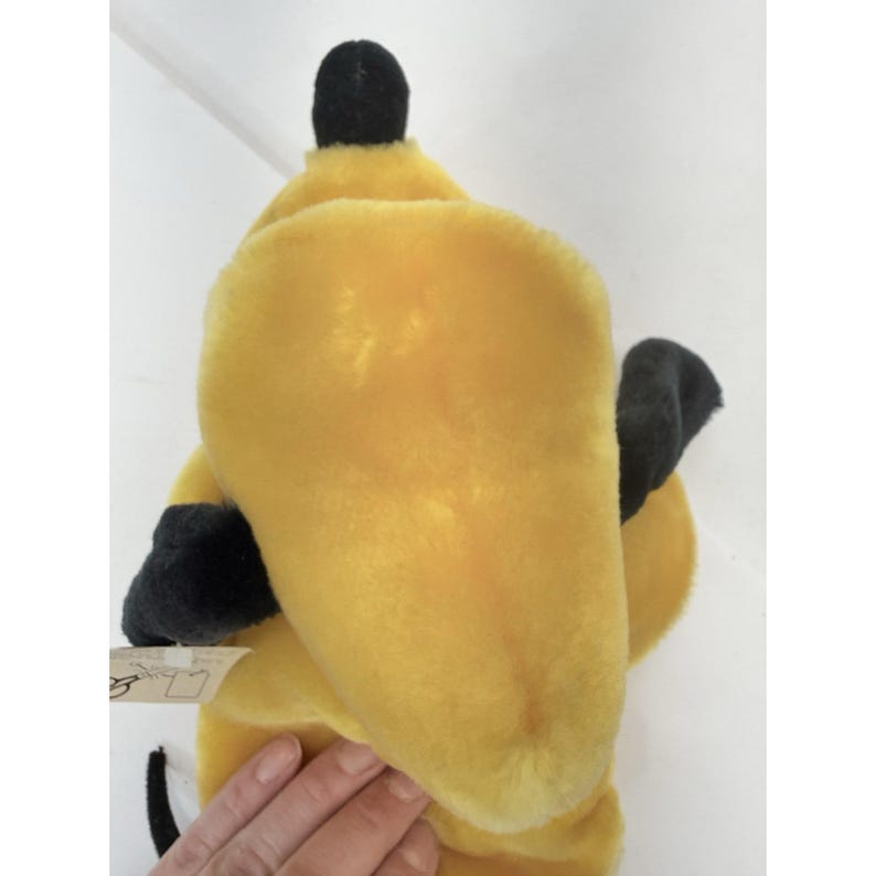 Puede incluir: Un juguete de peluche amarillo con detalles negros, incluyendo una parte superior y orejas negras. El juguete tiene una textura suave y una etiqueta adjunta. Es probable que sea un personaje de un cuento infantil o de dibujos animados.
