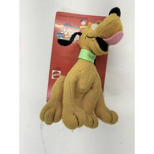 Puede incluir: Un peluche de Pluto, un perro amarillo con orejas negras y una nariz negra, con un collar verde. El juguete est&aacute; sujeto a una etiqueta de cart&oacute;n roja con el texto "FOR AGES OVER 18 MONTHS" y el logotipo de Mattel.