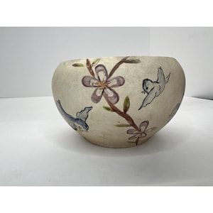 May include: A cream-colored, round ceramic bowl with hand-painted floral and bird designs. The flowers are purple and the birds are blue. The bowl has a wide opening and a smooth surface.
