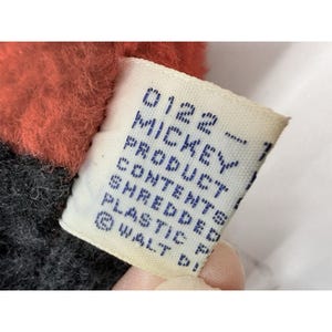 May include: Close-up of a tag from a Mickey Mouse product. The tag is white with blue text that reads "0122 - MICKEY PRODUCT CONTENTS SHREDDED PLASTIC &copy; WALT D!". The plush toy has red and black fabric.