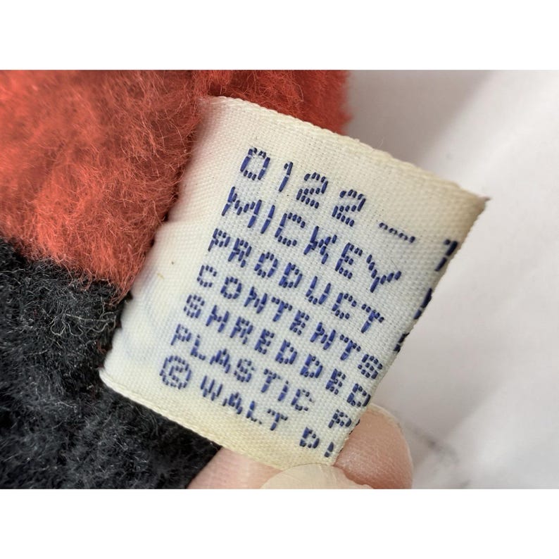 May include: Close-up of a tag from a Mickey Mouse product. The tag is white with blue text that reads "0122 - MICKEY PRODUCT CONTENTS SHREDDED PLASTIC &copy; WALT D!". The plush toy has red and black fabric.