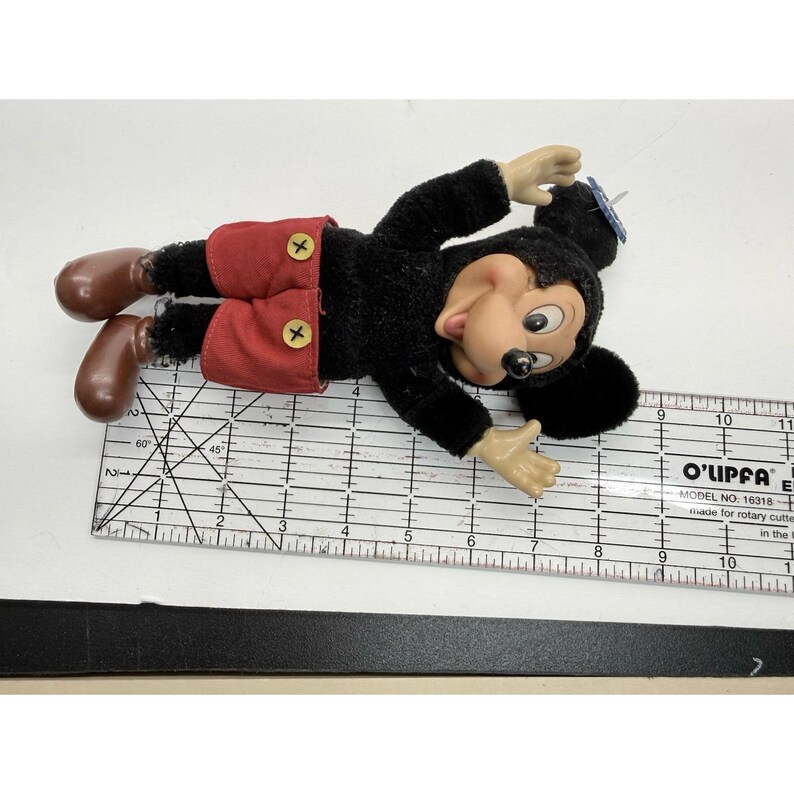 May include: A vintage Mickey Mouse plush doll, lying down. The doll has a black body, red shorts with yellow buttons, and brown shoes. The face is a light tan color with black eyes and a black nose. The doll is lying on a white surface.