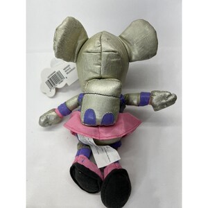 May include: A silver plush toy mouse with pink and purple accents. The mouse is wearing a pink skirt and has a purple backpack. The toy is sitting on a white surface.