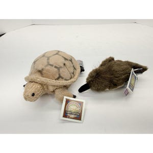 May include: Two plush animal toys: a desert tortoise and a platypus. The tortoise is tan with a patterned shell and the platypus is brown with a black bill and tail. Both have attached tags.