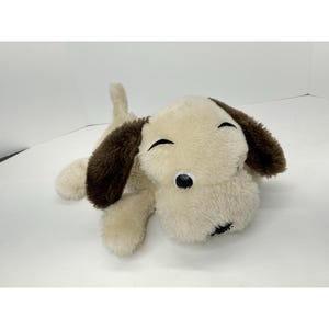 May include: A plush dog toy in a lying position. The toy has a cream-colored body with brown ears and a tail. It has black eyes and a black nose. The toy is made of soft, fuzzy material.