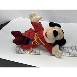 May include: A plush Mickey Mouse toy wearing a red robe with gold trim and a blue sash. The toy is lying on its back with its arms outstretched.