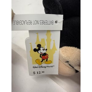 Peut inclure: Une peluche noire de Mickey Mouse avec un visage beige et une &eacute;tiquette indiquant "PILES NON REMPLA&Ccedil;ABLES". L'&eacute;tiquette pr&eacute;sente un dessin de Mickey Mouse et les mots "Walt Disney World".
