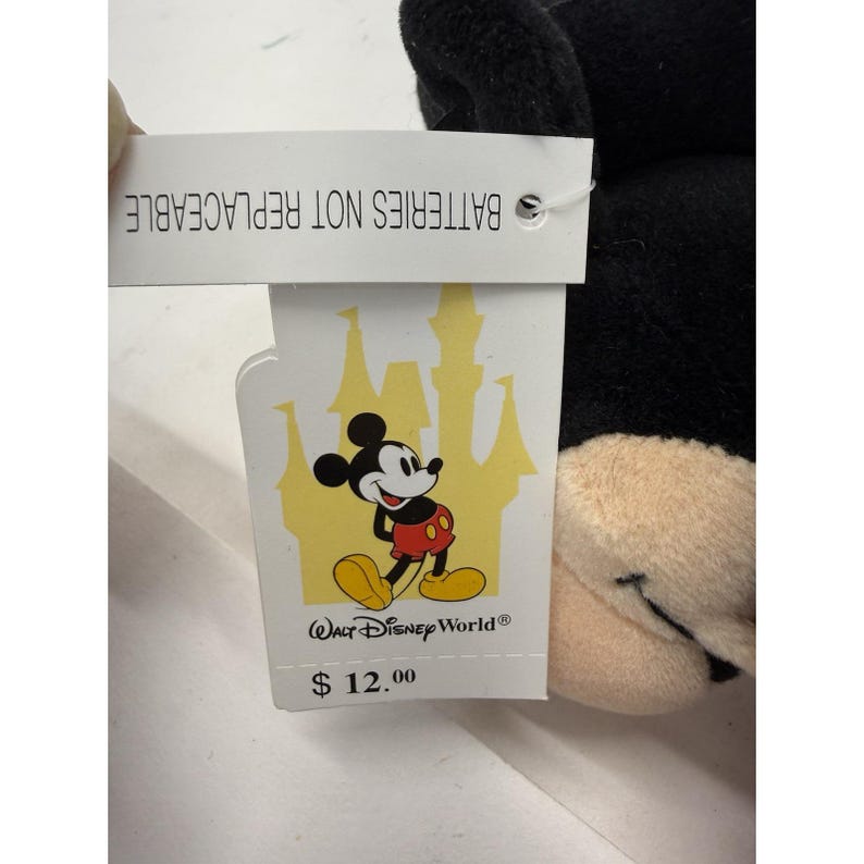 Peut inclure: Une peluche noire de Mickey Mouse avec un visage beige et une &eacute;tiquette indiquant "PILES NON REMPLA&Ccedil;ABLES". L'&eacute;tiquette pr&eacute;sente un dessin de Mickey Mouse et les mots "Walt Disney World".