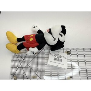 Puede incluir: Un peque&ntilde;o peluche de Mickey Mouse, tumbado de lado. El juguete presenta una cabeza y un cuerpo negros, pantalones cortos rojos con botones amarillos, guantes blancos y zapatos amarillos. Una etiqueta est&aacute; adjunta, con el texto "MINI BEANBAG 3D S MICKEY".
