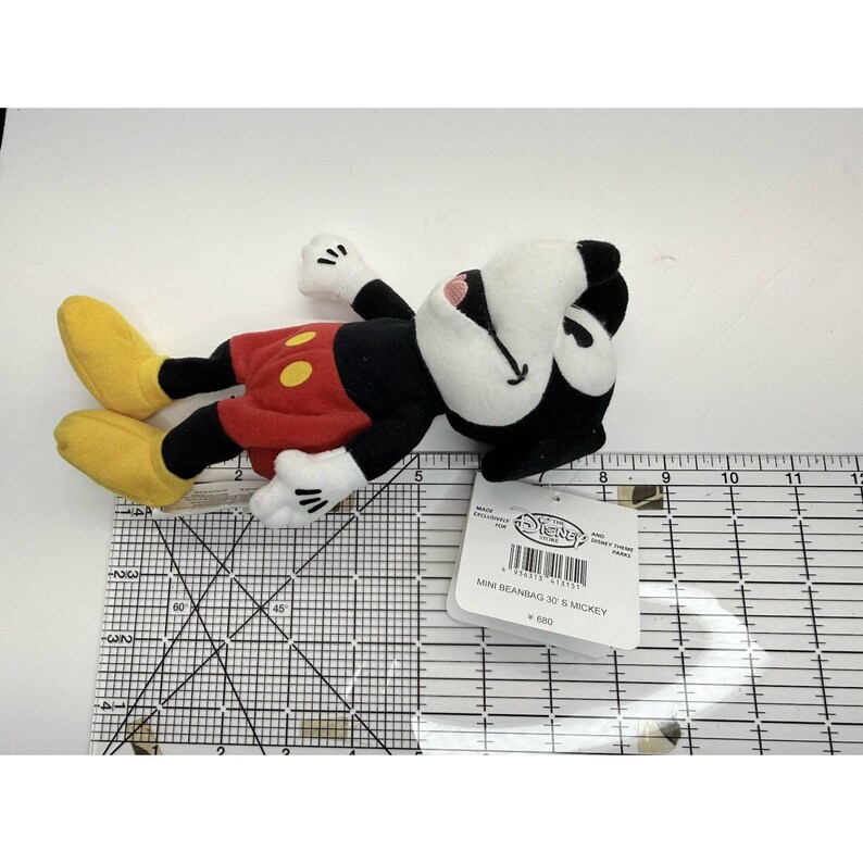 Puede incluir: Un peque&ntilde;o peluche de Mickey Mouse, tumbado de lado. El juguete presenta una cabeza y un cuerpo negros, pantalones cortos rojos con botones amarillos, guantes blancos y zapatos amarillos. Una etiqueta est&aacute; adjunta, con el texto "MINI BEANBAG 3D S MICKEY".