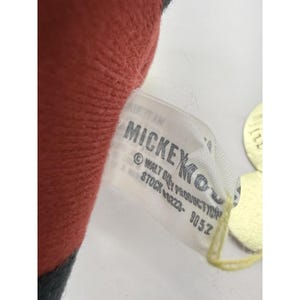 Puede incluir: Primer plano de la etiqueta de un peluche. La etiqueta es blanca con texto negro que dice "MICKEY" y "&copy; WALT DISNEY PRODUCTION". El juguete es rojo y negro.