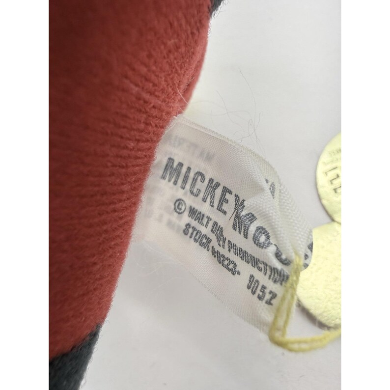 Puede incluir: Primer plano de la etiqueta de un peluche. La etiqueta es blanca con texto negro que dice "MICKEY" y "&copy; WALT DISNEY PRODUCTION". El juguete es rojo y negro.