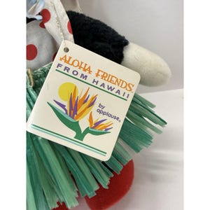 Puede incluir: Mu&ntilde;eca de peluche con cabeza negra, brazos blancos y pies rojos. Lleva una falda de hierba verde y un gorro blanco y rojo con lunares. La etiqueta dice "Aloha Friends From Hawaii by applause".