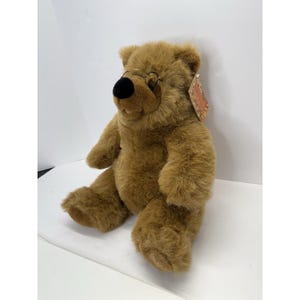 May include: A plush brown teddy bear with a black nose and a tag attached. The bear is sitting with its legs extended and arms at its sides. The fur is soft and textured.