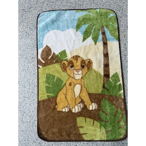May include: A brown, green, and blue plush blanket with a cartoon image of Simba from The Lion King. Simba is sitting on a brown rock with green leaves behind him. There is a palm tree in the background.