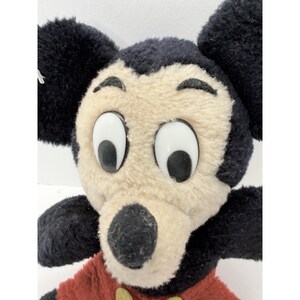 May include: Close-up of a vintage Mickey Mouse plush toy. The plush features a black head and ears, a tan face, and large white eyes. Mickey wears a red shirt with yellow buttons. A classic Disney collectible.
