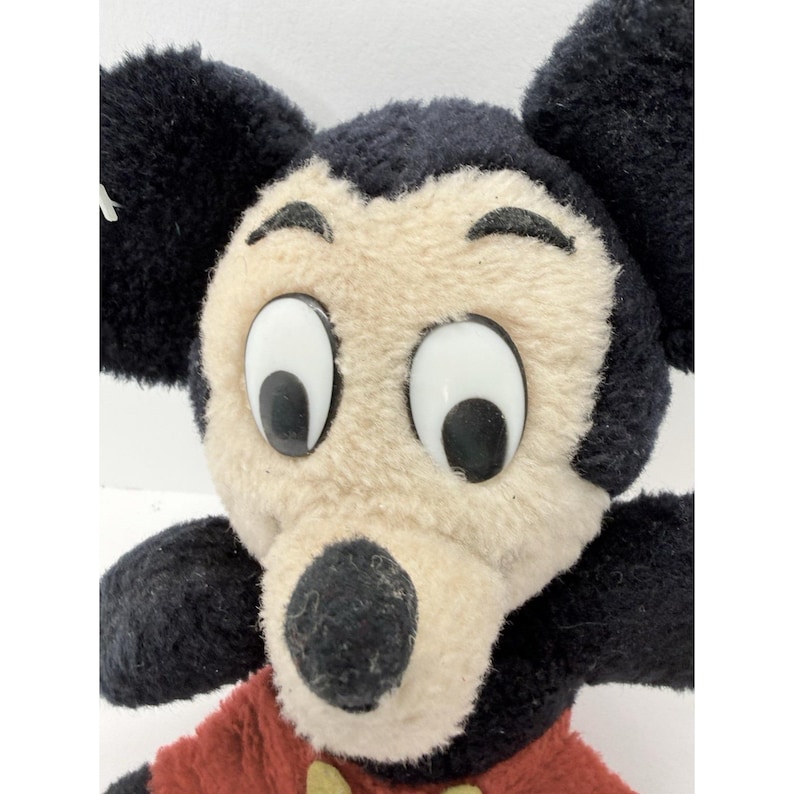 May include: Close-up of a vintage Mickey Mouse plush toy. The plush features a black head and ears, a tan face, and large white eyes. Mickey wears a red shirt with yellow buttons. A classic Disney collectible.