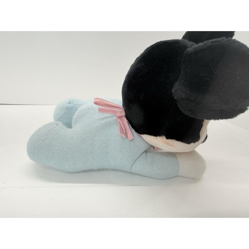 Puede incluir: Un peluche de un personaje en blanco y negro con grandes orejas negras. El personaje est&aacute; acostado boca abajo, con un atuendo azul claro y un lazo rosa. El juguete tiene detalles blancos en las manos y los pies.