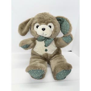 May include: A plush, brown teddy bear with long ears, a green bow tie, and a vest with white polka dots. The bear's ears and paws have a green pattern. The bear is sitting upright with its arms outstretched.