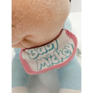Puede incluir: Un peluche con cuerpo azul claro y cabeza color melocot&oacute;n. El juguete tiene un babero con las palabras "Baby Mickey" en escritura azul claro, delineadas en rosa. El juguete es suave y parece estar hecho de un material de felpa.