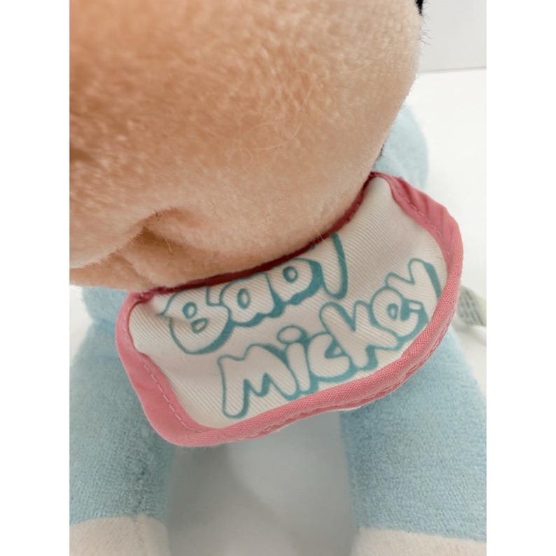 Puede incluir: Un peluche con cuerpo azul claro y cabeza color melocot&oacute;n. El juguete tiene un babero con las palabras "Baby Mickey" en escritura azul claro, delineadas en rosa. El juguete es suave y parece estar hecho de un material de felpa.