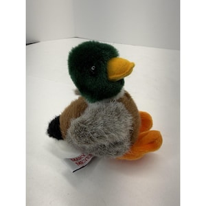 May include: A plush duck toy with a green head, yellow beak, and orange feet. The body features brown, gray, and black fur. A white tag with "MARY MEYER" is attached. This soft toy is suitable for children.