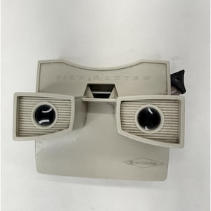 May include: A light gray plastic View-Master stereoscope viewer featuring two circular lenses, a horizontal ribbed design around the eye pieces, and a black advance lever on the side. The brand name View-Master and the manufacturer name Sawyer's are embossed.