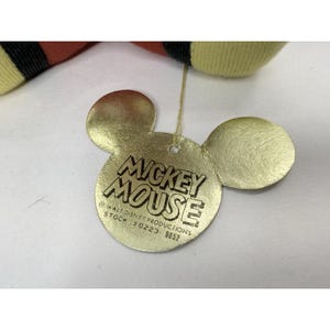 Puede incluir: Una etiqueta dorada de Mickey Mouse con las palabras "Mickey Mouse" en una fuente audaz y estilizada. La etiqueta tiene la forma de la cabeza y las orejas de Mickey Mouse. La etiqueta tambi&eacute;n incluye el texto "Walt Disney Productions" y un n&uacute;mero de stock.