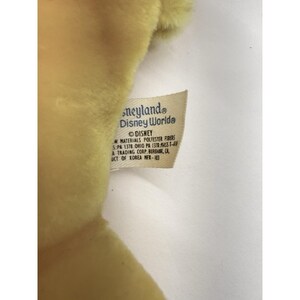 Puede incluir: Primer plano de un peluche amarillo con una etiqueta que dice "neyland Disney Worlde" y "DISNEY". La etiqueta tambi&eacute;n enumera los materiales como fibras de poli&eacute;ster y la ubicaci&oacute;n como Burbank, CA. El juguete est&aacute; hecho en Corea.