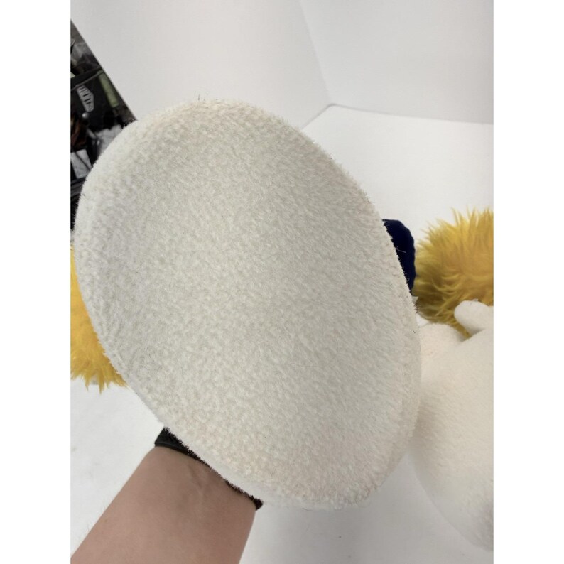 May include: Close-up of a large, white, plush slipper with a textured surface. The slipper has yellow and blue accents, and a white glove is visible. The slipper is part of a costume.