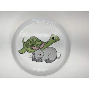 May include: White plastic plate featuring a cartoon illustration of a green turtle with a spotted shell and a gray rabbit with pink ears. The plate has a simple, clean design.