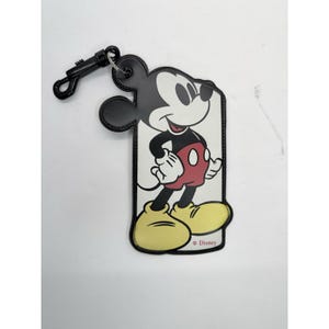 May include: A Mickey Mouse luggage tag with a black border and a black clip. The tag features a full-body image of Mickey Mouse in his classic outfit: red shorts, yellow shoes, and white gloves. The Disney logo is visible.
