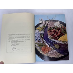 May include: An open cookbook with a colorful food arrangement on the right page. The arrangement features various foods and flowers, arranged in a decorative manner. The left page contains text.