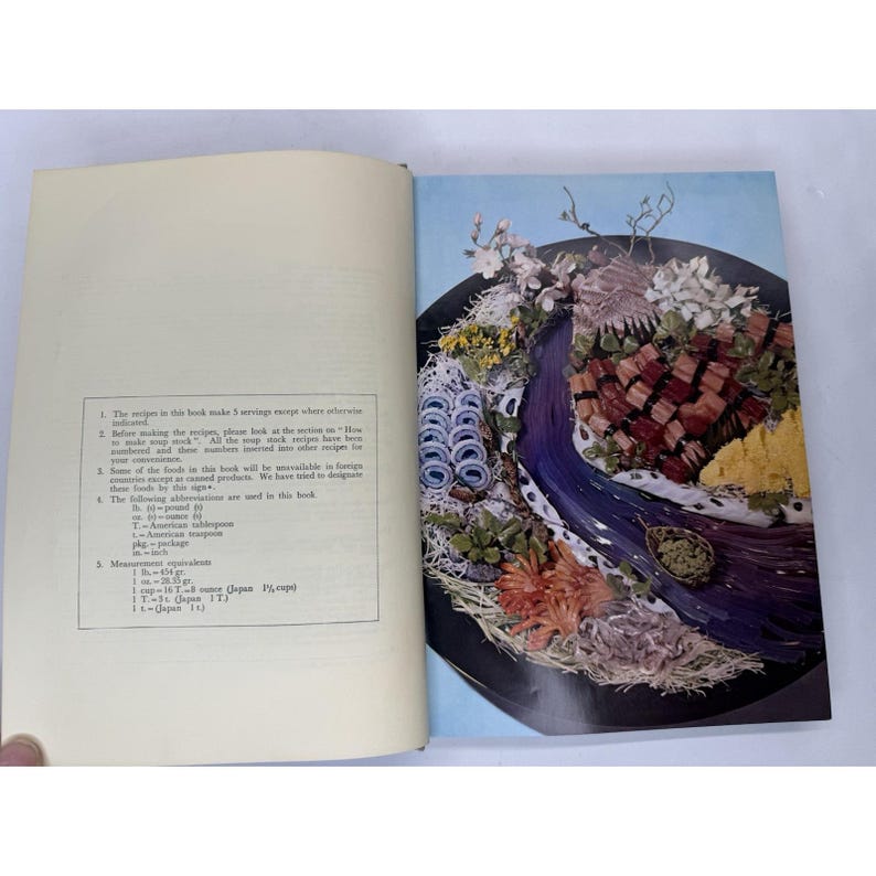 May include: An open cookbook with a colorful food arrangement on the right page. The arrangement features various foods and flowers, arranged in a decorative manner. The left page contains text.
