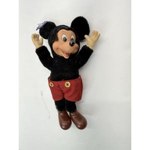 May include: A vintage Mickey Mouse plush doll. The doll has a black body, head, and ears, with a tan face and hands. It wears red shorts with yellow buttons and brown shoes. The doll's arms are raised in a welcoming gesture.