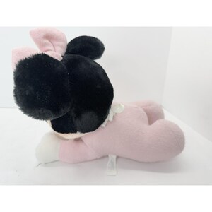 Puede incluir: Un mu&ntilde;eco de peluche de Minnie Mouse en posici&oacute;n acostada. El mu&ntilde;eco tiene una cabeza negra con orejas grandes y redondas y un lazo rosa. El cuerpo es rosa, con guantes blancos. El mu&ntilde;eco est&aacute; hecho de material suave y afelpado.
