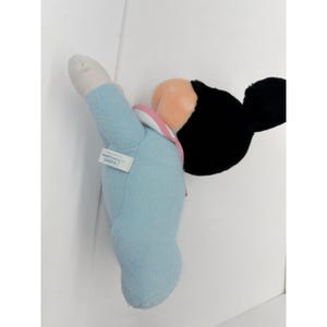 Puede incluir: Un mu&ntilde;eco de peluche de Mickey Mouse con un atuendo azul claro y un cuello rosa. El mu&ntilde;eco tiene orejas negras, una cara color melocot&oacute;n y una mano blanca. Una peque&ntilde;a etiqueta est&aacute; adherida al brazo del mu&ntilde;eco.