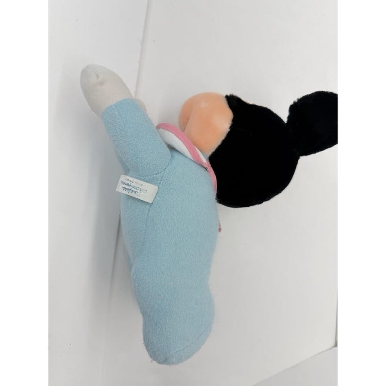 Puede incluir: Un mu&ntilde;eco de peluche de Mickey Mouse con un atuendo azul claro y un cuello rosa. El mu&ntilde;eco tiene orejas negras, una cara color melocot&oacute;n y una mano blanca. Una peque&ntilde;a etiqueta est&aacute; adherida al brazo del mu&ntilde;eco.