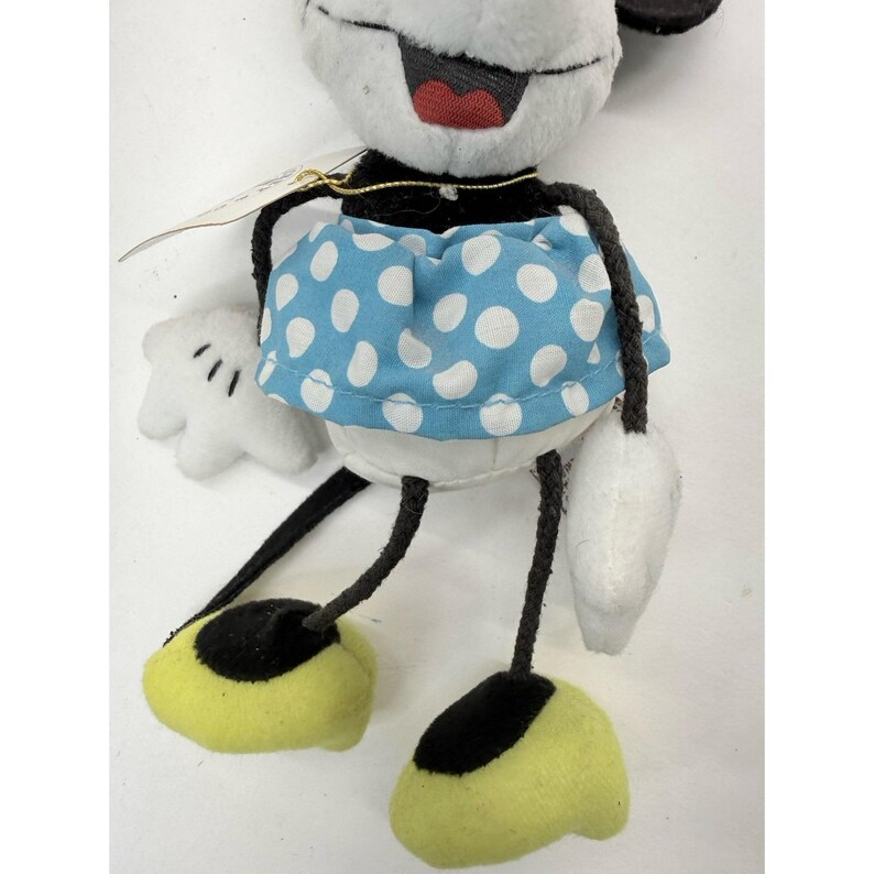 K&ouml;nnte beinhalten: Pl&uuml;schpuppe Minnie Mouse mit wei&szlig;em Gesicht, schwarzen Ohren und einem blauen Polka-Dot-Rock. Die Puppe hat schwarze Beine und Arme, gelbe F&uuml;&szlig;e mit schwarzen Sohlen und wei&szlig;e Handschuhe. Ein goldenes Etikett ist angebracht.